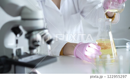 A scientist, wearing a lab coat and pink protective gloves, is pouring a yellow oily liquid from one beaker to another near microscope in laboratory, close up. Medicine and science 122955297