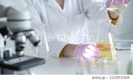 A scientist, wearing a lab coat and pink protective gloves, is pouring a yellow oily liquid from one beaker to another near microscope in laboratory, close up. Medicine and science A scientist, wearing a lab coat and pink protective gloves, is pouring a yellow oily liquid from one beaker to another near microscope in laboratory, close up. Medicine and science 122955300