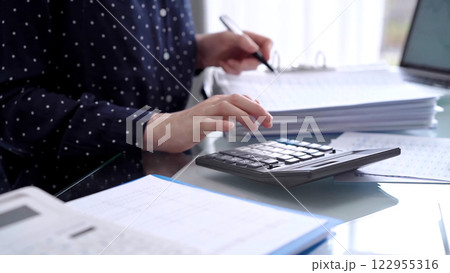 Businesswoman using calculator and reviewing ring folder of financial documents with magnifying glass at desk in modern office. Audit and taxes in business Businesswoman using calculator and reviewing ring folder of financial documents with magnifying glass at desk in modern office. Audit and taxes in business 122955316