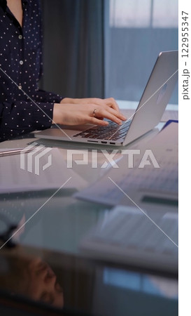 Business woman with dotted blues typing on laptop computer while sitting at the glass table in office in the evening, closeup 122955347