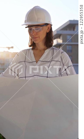 Woman an architect, wearing safety helmets and glasses, is holding blueprints and inspecting a building site during sunrise or sunset, front view vertical portrait. Architecture concept Woman an architect, wearing safety helmets and glasses, is holding blueprints and inspecting a building site during sunrise or sunset, front view vertical portrait. Architecture concept 122955358