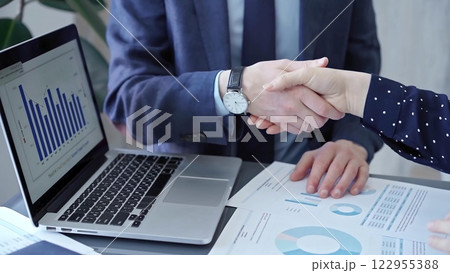 Two business people professionals shaking hands in office setting, sealing a deal with laptop and financial graphs visible. Business handshake over laptop and reports 122955388
