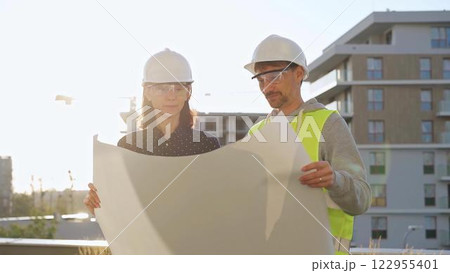 Two architects wearing safety hard hats are holding blueprint and discussing something on construction site at sunrise. Architect and teamwork concept 122955401
