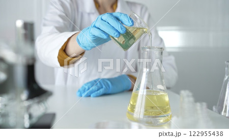 A scientist, wearing a lab coat and blue protective gloves, is pouring a yellow liquid from one beaker to another in laboratory, close up. Medicine and science concepts 122955438