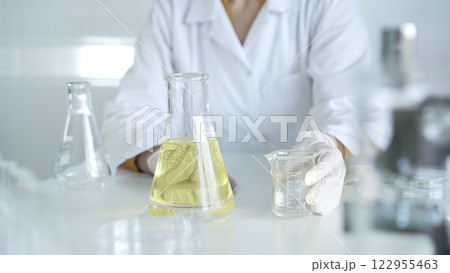 A scientist, wearing a lab coat and white protective gloves, is holding a yellow oily liquid inside beaker in laboratory setting, close up. Medicine and science concept 122955463
