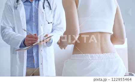 Doctor man is making notes on a clipboard while patient woman is describing back pain. Medicine and orthopedics concepts Doctor man is making notes on a clipboard while patient woman is describing back pain. Medicine and orthopedics concepts 122955464