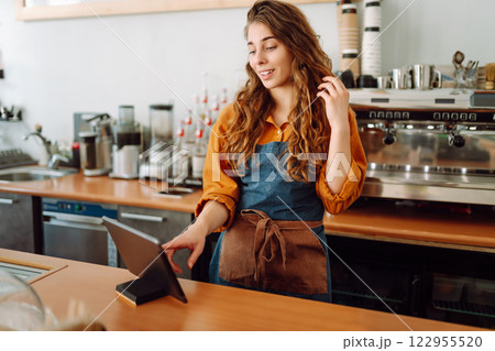 Smiling female barista takes an order from a tablet while standing at bar counter in a coffee shop. Smiling female barista takes an order from a tablet while standing at bar counter in a coffee shop. 122955520