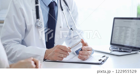 Doctor and a patient. The physician, wearing a white medical coat over a blue shirt and tie, gesturing with his hands during a consultation in the clinic. Medicine concept Doctor and a patient. The physician, wearing a white medical coat over a blue shirt and tie, gesturing with his hands during a consultation in the clinic. Medicine concept 122955529
