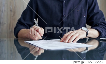 Businessman in dark blue t-shirt is signing contract at desk. Close-up of a male executive signing a legal document on a glass desk. Business people concept Businessman in dark blue t-shirt is signing contract at desk. Close-up of a male executive signing a legal document on a glass desk. Business people concept 122955542
