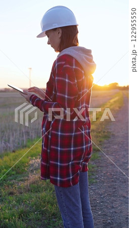 Woman engineer wearing a white protective helmet is taking notes with a clipboard in a field with wind turbines, as the sun sets. Clean energy and engineering concept 122955550