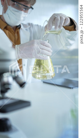 Man scientist wearing a lab coat, white gloves, mask, and protective glasses, is pouring a yellow oily liquid from one beaker to another near microscope in a laboratory setting, close up. Science and Man scientist wearing a lab coat, white gloves, mask, and protective glasses, is pouring a yellow oily liquid from one beaker to another near microscope in a laboratory setting, close up. Science and 122955564