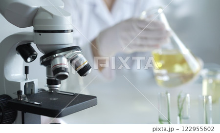 Microscope is near a scientist, wearing a lab coat and white protective gloves, and holding beaker with a yellow oily liquid in laboratory setting, close up. Medicine and science concept 122955602