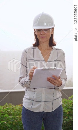 Female construction engineer wearing white checked blouse and safety helmet is making notes on a clipboard while inspecting a building site in foggy weather, front vertical view. Architecture concept 122955605
