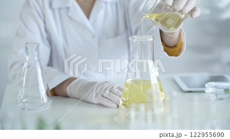 A scientist, wearing a lab coat and white protective gloves, is pouring a yellow oily liquid from one beaker to another in laboratory, close up. Medicine and science concept A scientist, wearing a lab coat and white protective gloves, is pouring a yellow oily liquid from one beaker to another in laboratory, close up. Medicine and science concept 122955690