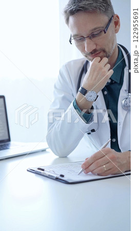 Doctor and a patient. The physician, wearing a white medical coat over a green shirt, gesturing with his hands during a consultation in the clinic. Medicine concept 122955691