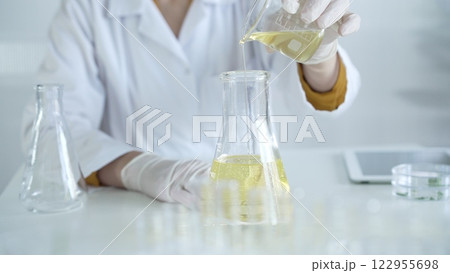 A scientist, wearing a lab coat and white protective gloves, is pouring a yellow oily liquid from one beaker to another in laboratory, close up. Medicine and science concept A scientist, wearing a lab coat and white protective gloves, is pouring a yellow oily liquid from one beaker to another in laboratory, close up. Medicine and science concept 122955698