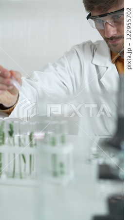 A male science researcher, wearing a lab coat and protective glasses, drips liquid from a pipette into a test tube containing a green plant. His face is focused, captured in a vertical portrait A male science researcher, wearing a lab coat and protective glasses, drips liquid from a pipette into a test tube containing a green plant. His face is focused, captured in a vertical portrait 122955702