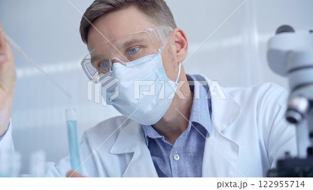 Man with safety glasses and blue mask is working with lab tubes using a pipette in a modern laboratory, close-up of researcher. Science and medicine Man with safety glasses and blue mask is working with lab tubes using a pipette in a modern laboratory, close-up of researcher. Science and medicine 122955714