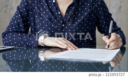 Businesswoman is signing corporate contract at her office desk in formal dark blue blouse with polka dots. Business people concept Businesswoman is signing corporate contract at her office desk in formal dark blue blouse with polka dots. Business people concept 122955727