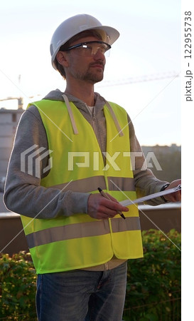 Man engineer with white helmet and safety vest is taking notes on a clipboard while inspecting a construction site at sunrise in early morning, vertical front view. Architecture and engineering Man engineer with white helmet and safety vest is taking notes on a clipboard while inspecting a construction site at sunrise in early morning, vertical front view. Architecture and engineering 122955738