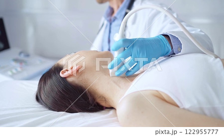 Doctor man wearing blue medical gloves is using ultrasound equipment on neck of female patient lying down for a medical examination 122955777