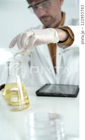 Man scientist with lab coat, white gloves, and protective glasses, is pouring a yellow oily liquid from one beaker to another near microscope in a laboratory, vertical close-up view 122955781