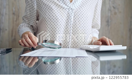 Auditor woman analyzing financial documents with magnifying glass and calculator at her office desk in formal white blouse with polka dots. Business people concept 122955783