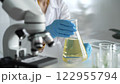 Scientist, wearing blue protective gloves, is holding a beaker with yellow liquid while sitting at the table in laboratory besides microscope, close up. Medicine and science concepts 122955794