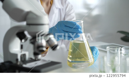 Scientist, wearing blue protective gloves, is holding a beaker with yellow liquid while sitting at the table in laboratory besides microscope, close up. Medicine and science concepts 122955794