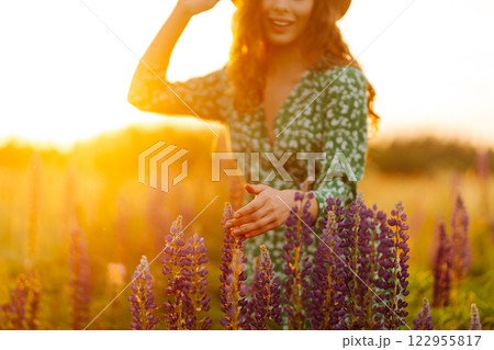Close-up of young woman in hat and curly hair touching lupins in field at sunset. Fashion and nature Close-up of young woman in hat and curly hair touching lupins in field at sunset. Fashion and nature 122955817