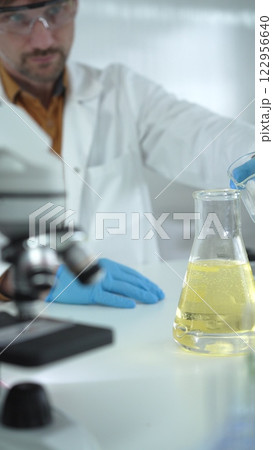 Man scientist with lab coat, blue gloves, and protective glasses, is pouring a yellow oily liquid from one beaker to another near microscope in a laboratory, vertical close-up view Man scientist with lab coat, blue gloves, and protective glasses, is pouring a yellow oily liquid from one beaker to another near microscope in a laboratory, vertical close-up view 122956640