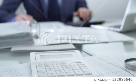 Magnifying glass, calculator, pen, laptop and documents are lying on accountant's desk. Businessman wearing blue suit is working with documents on the background. Business people and audit concept Magnifying glass, calculator, pen, laptop and documents are lying on accountant's desk. Businessman wearing blue suit is working with documents on the background. Business people and audit concept 122956646