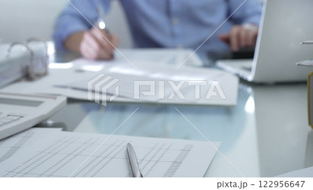 Calculator, pen and documents are lying on accountant's desk. Businessman wearing blue shirt is working with documents on the background. Business people, audit concept Calculator, pen and documents are lying on accountant's desk. Businessman wearing blue shirt is working with documents on the background. Business people, audit concept 122956647