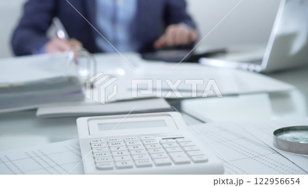 Magnifying glass, calculator, pen, laptop and documents are lying on accountant's desk. Businessman wearing blue suit is working with documents on the background. Business people and audit concept Magnifying glass, calculator, pen, laptop and documents are lying on accountant's desk. Businessman wearing blue suit is working with documents on the background. Business people and audit concept 122956654