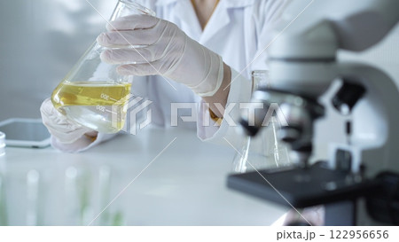 Microscope is near a scientist, wearing a lab coat and white protective gloves, and holding beaker with a yellow oily liquid in laboratory setting, close up. Medicine and science concept 122956656