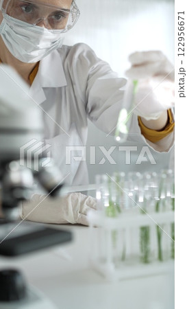 Woman scientist wearing lab coat, white gloves, face mask and protective glasses, is holding a test tube with plants inside near microscope in laboratory, vertical portrait view. Science and medicine 122956671