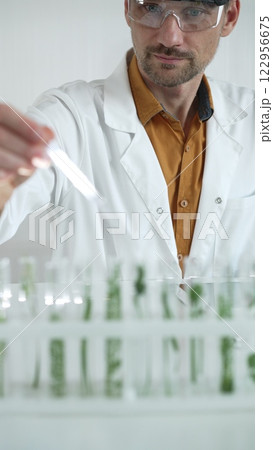 A male science researcher, wearing a lab coat and protective glasses, drips liquid from a pipette into a test tube containing a green plant. His face is focused, captured in a vertical portrait 122956675