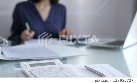 White calculator, pen and documents are lying on accountant's desk. Businesswoman wearing blue dress is working with documents on the background. Business and audit concept White calculator, pen and documents are lying on accountant's desk. Businesswoman wearing blue dress is working with documents on the background. Business and audit concept 122956727