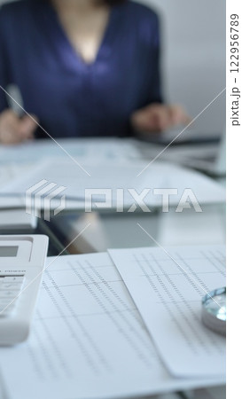 Businesswoman wearing blue dress is working with laptop, calculator and documents in a professional office setting. Low lighting, close-up vertical view. Business people, audit concept Businesswoman wearing blue dress is working with laptop, calculator and documents in a professional office setting. Low lighting, close-up vertical view. Business people, audit concept 122956789