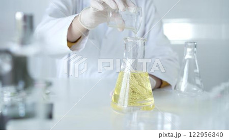 A scientist, wearing a lab coat and white protective gloves, is pouring a yellow oily liquid from one beaker to another in laboratory, close up. Medicine and science concept 122956840