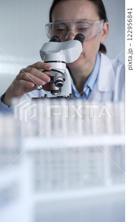 Woman scientist wearing lab coat and protective glasses is using microscope in modern laboratory setup. Low key lighting photo. Science and Medicine concepts 122956841
