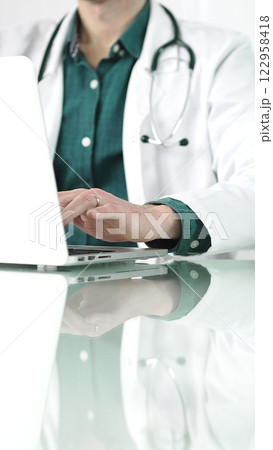 Doctor man in a green shirt under a medical coat is using technology in medicine. Close-up of hands typing on a laptop on a glass desk in the clinic, vertical view. Healthcare concept 122958418