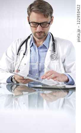 Doctor man in blue shirt under a white coat, glasses and stethoscope is using digital tablet in modern healthcare setting, interacting with patient data, vertical portrait view. Medicine and science 122958422