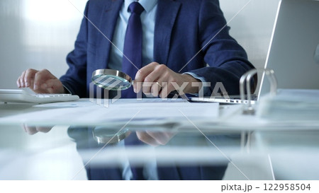 Businessman analyzing financial data with magnifying glass and calculator, conducting audit or review in office. Audit and taxes theme in business Businessman analyzing financial data with magnifying glass and calculator, conducting audit or review in office. Audit and taxes theme in business 122958504