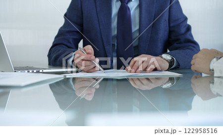 Businessman pointing at charts with pen, explaining data to colleague while sitting at the glass table in office. Businesspeople concept Businessman pointing at charts with pen, explaining data to colleague while sitting at the glass table in office. Businesspeople concept 122958516