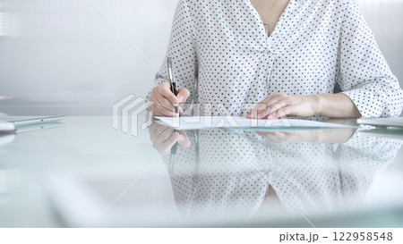 Businesswoman in white dotted blouse is signing a contract at a sleek glass desk in a modern office, showcasing professionalism and focus in her work environment. Business people concept Businesswoman in white dotted blouse is signing a contract at a sleek glass desk in a modern office, showcasing professionalism and focus in her work environment. Business people concept 122958548