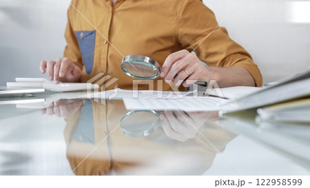 Businessman accountant wearing casual yellow shirt carefully reviewing financial reports with magnifying glass and calculator, conducting audit and analyzing data in office. Taxes, audit in business 122958599