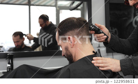 Skilled barber trimming client's hair using electric razor, creating contemporary hairstyle in modern barbershop setting with precise, professional technique Skilled barber trimming client's hair using electric razor, creating contemporary hairstyle in modern barbershop setting with precise, professional technique 122958611