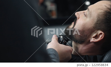 Skilled barber grooming client's beard using electric razor, delivering precise styling within contemporary barbershop setting Skilled barber grooming client's beard using electric razor, delivering precise styling within contemporary barbershop setting 122958634