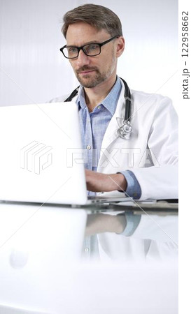 Doctor man in a blue shirt under a medical coat is using technology in medicine. Portrait of physician typing on a laptop on a glass desk in clinic, vertical view. Medicine and science 122958662